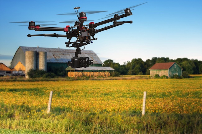 A flying helicopter with raised landing gears and a camera with blured crop field and farm structures on a background highlighted by a sunset