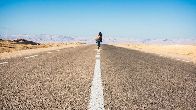 Woman walking down middle of highway with a few belongings.